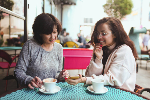 Mature mother and her young daughter sit together in cafe or restaurant. Girl hold smartphone and showing something on screen to her mom. Mature woman watch it. Drinking tea or coffee. - Fotoğraf, Görsel