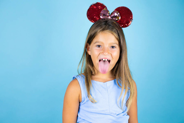 Mladý krásný dítě dívka nosí uši myši přes izolované modré pozadí vyčnívající jazyk velmi šťastný - Fotografie, Obrázek