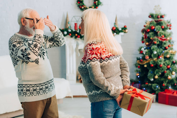 senior man covering eyes with hands while wife holding present behind back - Foto, afbeelding