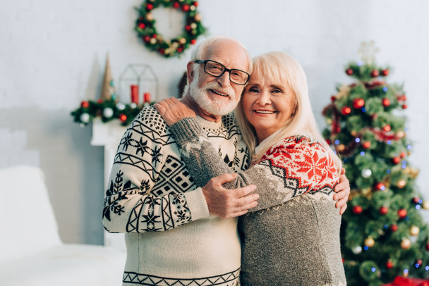 背景にクリスマスの装飾が施されたカメラを見ながら抱きしめる幸せなシニアカップル - 写真・画像