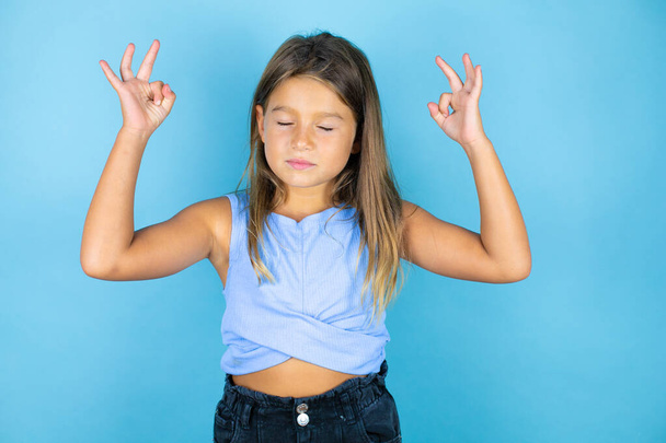 jong mooi kind meisje over geïsoleerde blauwe achtergrond ontspannen en glimlachen met de ogen gesloten doen meditatie gebaar met vingers - Foto, afbeelding