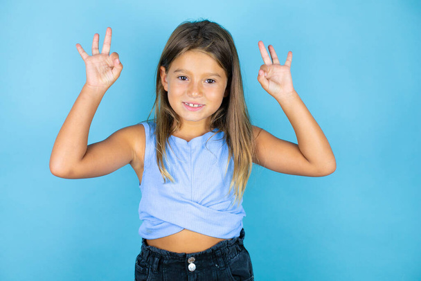 Mladý krásný dítě dívka přes izolované modré pozadí dělá v pořádku znamení s prsty a usmívá se, vynikající symbol - Fotografie, Obrázek