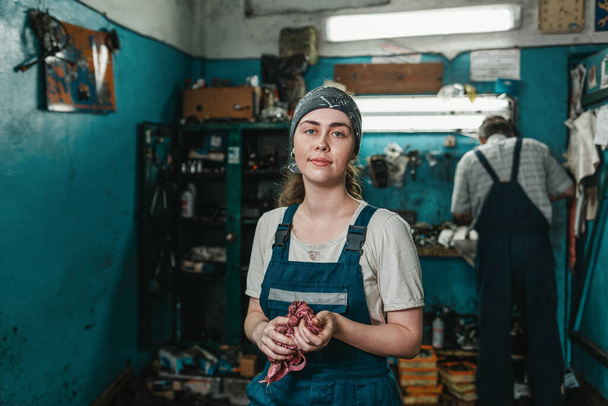Gender equality. Portrait of a young smiling woman in uniform working in a workshop, who wipes her hands with a rag. In the background, a worker at the machine. - Photo, Image