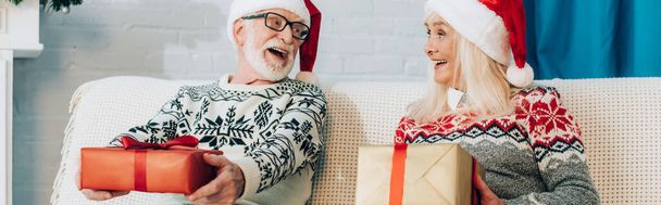 ιστοσελίδα κεφαλίδα ενθουσιασμένοι ηλικιωμένοι ζευγάρι σε Σάντα καπέλα κοιτάζοντας ο ένας τον άλλο, ενώ κάθεται στον καναπέ με κουτιά δώρων - Φωτογραφία, εικόνα