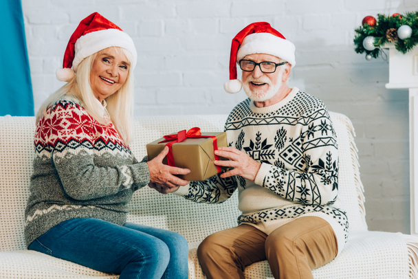 onnellinen vanhempi pari joulupukki hatut katsomassa kameraa istuessaan sohvalla lahjapakkauksella - Valokuva, kuva