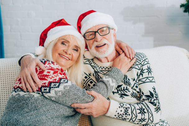 happy senior couple in santa hats embracing while looking at camera - Foto, afbeelding