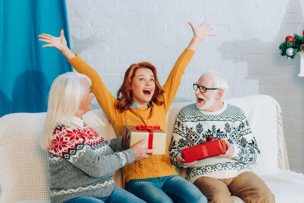 excited woman with raised hands sitting on sofa near senior parents holding christmas gifts - Foto, afbeelding