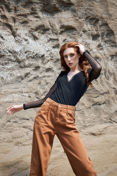 Fashion red-Haired girl in orange jeans posing in nature near sandy rocks, not like everyone else - Foto, Imagen