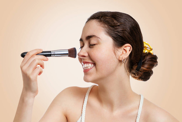 Retrato de una joven mujer feliz con una sonrisa aplicando maquillaje con un pincel. Fondo beige. Concepto de cosmetología, cosmética y cuidado de la piel. - Foto, imagen