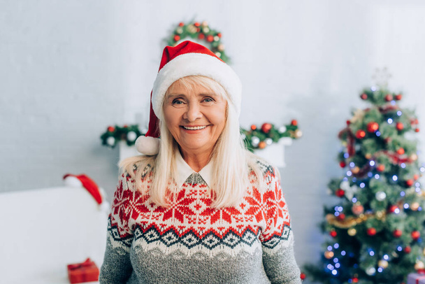 smiling senior woman in santa hat looking at camera near christmas tree on background - Photo, Image