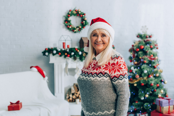 χαρούμενη ηλικιωμένη γυναίκα στο καπέλο santa κοιτάζοντας κάμερα κοντά σε χριστουγεννιάτικη διακόσμηση στο παρασκήνιο - Φωτογραφία, εικόνα