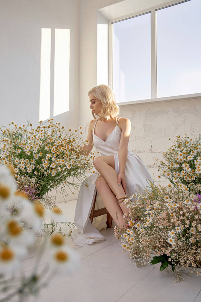 Sexy blonde woman in a beautiful white dress is sitting near the window in front of a bouquet of wild flowers. Romantic girl with beautiful natural makeup - Фото, изображение