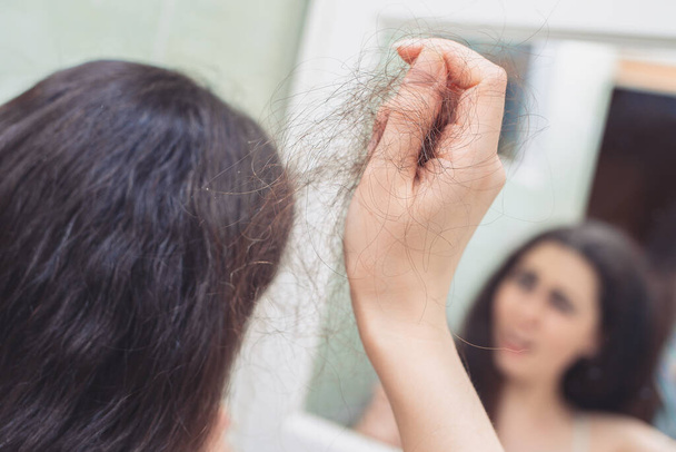 A woman pulls a bunch of hair from her head. Rear view. In the background, a reflection in the mirror. The concept of hair loss. - Foto, afbeelding