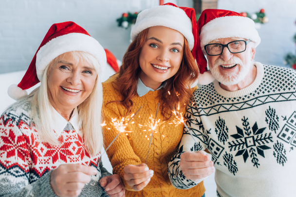 fröhliches Seniorenpaar mit Tochter in Weihnachtsmannmütze, Wunderkerzen in der Hand, während sie in die Kamera schauen - Foto, Bild