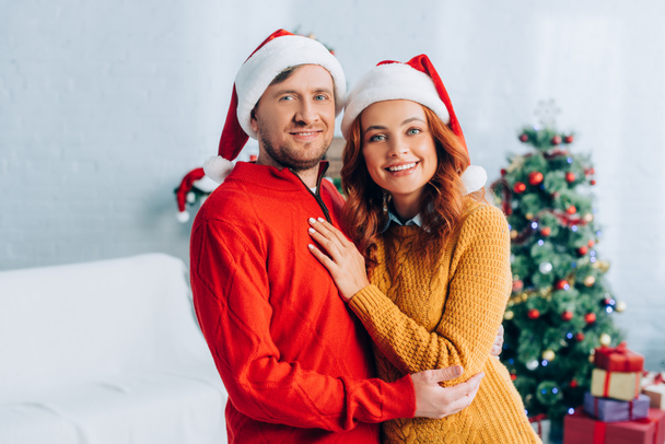 happy young couple in santa hats embracing while looking at camera - Φωτογραφία, εικόνα