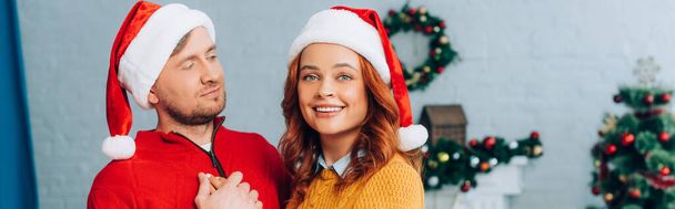 horizontal image of woman in santa hat smiling at camera while embracing husband on christmas - 写真・画像