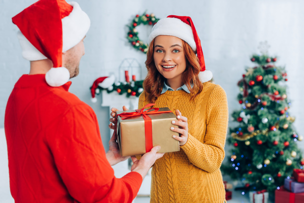 selective focus of excited woman in santa hat taking christmas gift from husband - Photo, Image