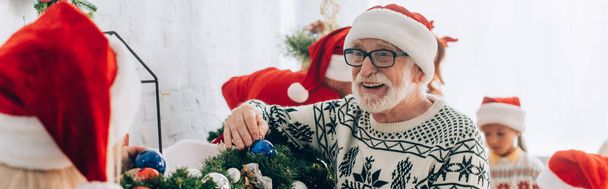 панорамний знімок щасливого старшого чоловіка в капелюсі Санта, що прикрашає гілку сосни разом з сім'єю
 - Фото, зображення