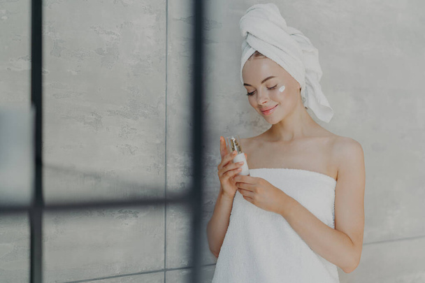 Satisfait jeune femme caucasienne prend soin de sa beauté, applique crème lotion sur le visage, se tient enveloppé dans une serviette de bain, pose dans la salle de bain à la maison contre le mur gris. Soins quotidiens à domicile et concept de spa de jour - Photo, image
