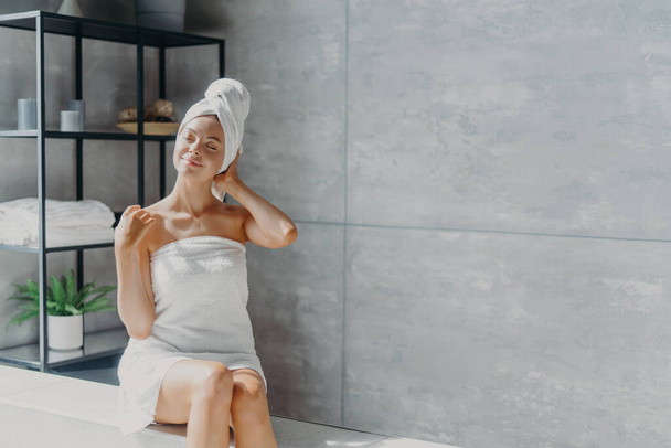 Relaxed young Caucasian female model wears towel wrapped on head, feels refreshed after taking shower, has healthy clean soft skin, poses in cozy bathroom. Women, beauty and hygiene concept. - Photo, image