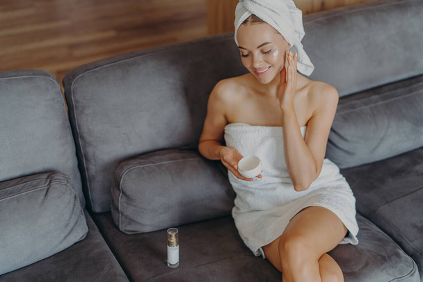 Η κορυφαία άποψη της ικανοποιημένης υγιούς γυναίκας κάθεται σε άνετο καναπέ εσωτερικό, εφαρμόζει κρέμα προσώπου και χρησιμοποιεί λοσιόν σώματος μετά το μπάνιο, έχει φυσικό μακιγιάζ και υγιές δέρμα. Έννοια υγιεινής και ομορφιάς - Φωτογραφία, εικόνα