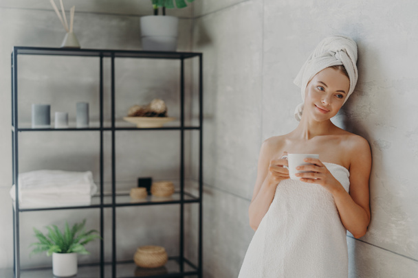 Photo of calm relaxed woman poses in towel against bathroom interior, wrapped in bath towel, drinks tea or coffee, has clean perfect skin, leans at grey wall. Beauty day, spa and skin care treatment - Photo, Image