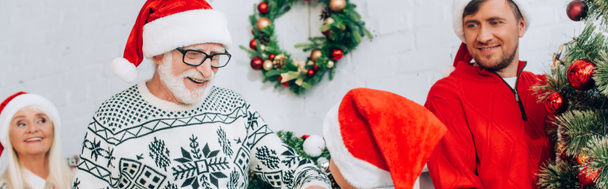 concetto panoramico di uomo anziano in cappello di Babbo Natale guardando nipote vicino alla famiglia e albero di Natale - Foto, immagini