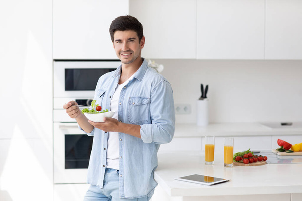 Handsome smiling young man with plate of salad fresh vegetables looking at camera. Portrait of happy casual guy in the kitchen. - Foto, immagini