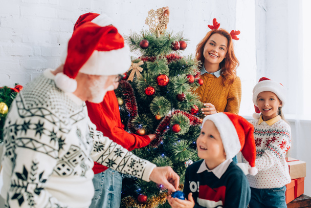 szelektív fókusz nagyapa ad csecsebecse unokája, miközben díszíti karácsonyfa együtt a család - Fotó, kép