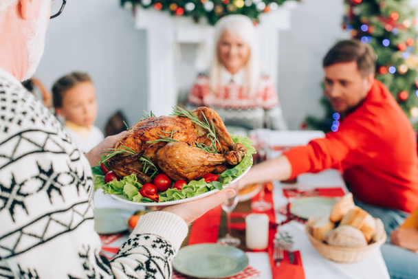 selective focus of senior man holding plate with traditional roasted turkey near family sitting at festive table - Foto, Imagen