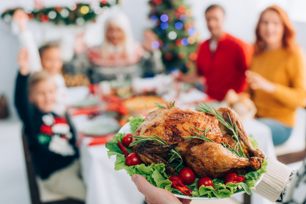 selective focus of senior man holding plate with traditional roasted turkey, cherry tomatoes and lettuce during christmas dinner with family - Фото, изображение