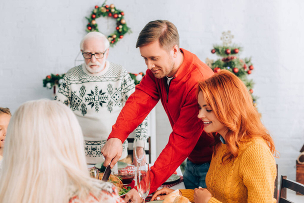 Uomo sorridente che taglia tacchino sul tavolo festivo con cena vicino alla famiglia a casa - Foto, immagini