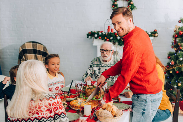 Sonriente hombre mirando a la cámara mientras corta pastel en la mesa festiva cerca de la familia - Foto, imagen