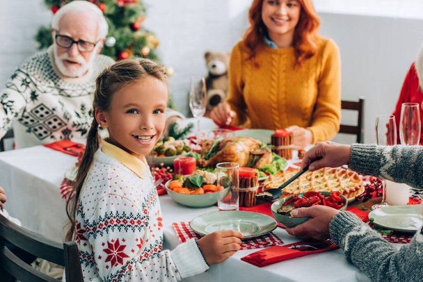Selective focus of girl looking at camera, sitting at festive table near family - Foto, Imagem