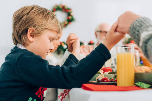 Επιλεκτική εστίαση του νηπιαγωγείου αγόρι κρατώντας τα χέρια με την οικογένεια, ενώ κάθεται στο τραπέζι - Φωτογραφία, εικόνα