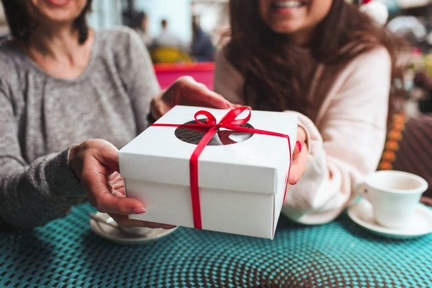 Mature mother and her young daughter sit together in cafe or restaurant. Close up and cut view of women holding one present together. White box tied up with red stripe. Christmas time or new year. - 写真・画像