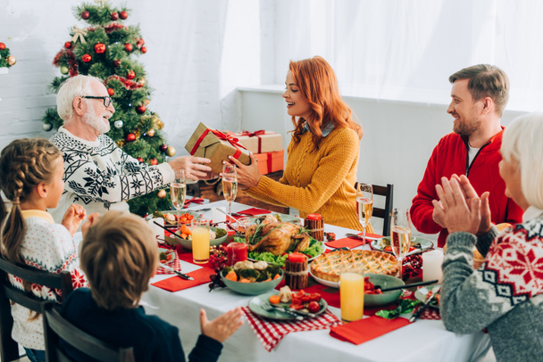Redhead woman greeting senior man with gift box, sitting at table with family - Photo, Image
