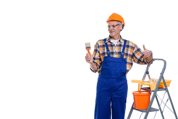 Professional builder, senior man in coverall and helmet ready for painting wall with roller, stay near stepladder, copy space and place for text. - Фото, зображення