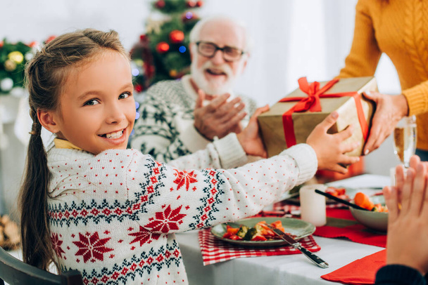 Selective focus of smiling girl with present from mother, sitting at festive table - Photo, Image