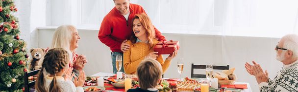 Панорамный снимок смеющейся женщины с подарком от мужчины, сидящей с семьей - Фото, изображение