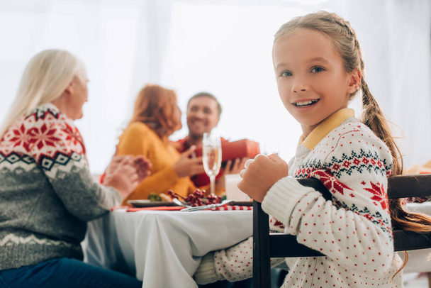 Selectieve focus van lachend meisje kijkend naar camera, zittend aan tafel met familie - Foto, afbeelding