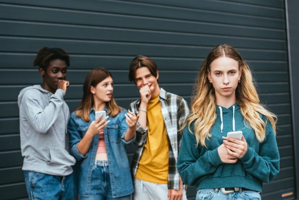 Enfoque selectivo de la chica con teléfono inteligente de pie cerca riendo adolescentes multiculturales que señalan con el dedo al aire libre  - Foto, Imagen