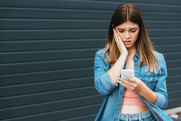 Worried teenager using smartphone near building outdoors  - Photo, Image