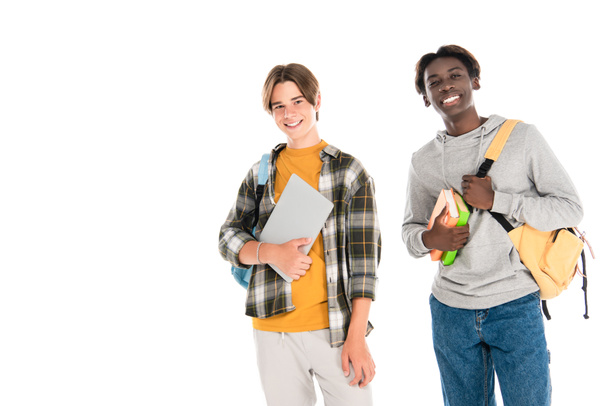 Lächelnde multiethnische Teenager mit Laptop und Büchern, die isoliert auf weiß in die Kamera schauen - Foto, Bild
