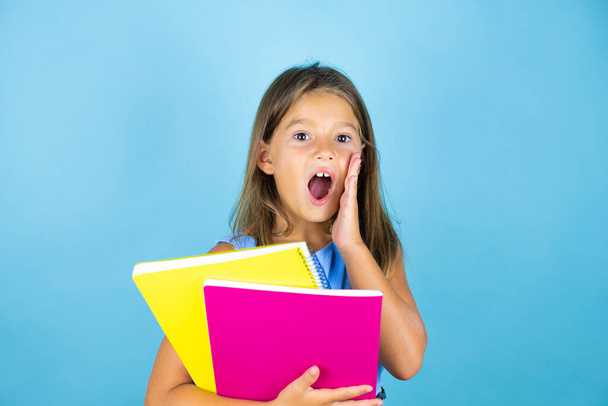 Young beautiful student child girl holding notebook over isolated blue background shouting and screaming loud to side with hands on mouth - Foto, Imagen