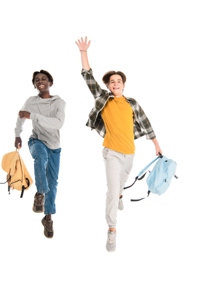 Cheerful multiethnic teenagers with backpacks jumping isolated on white - Foto, Bild