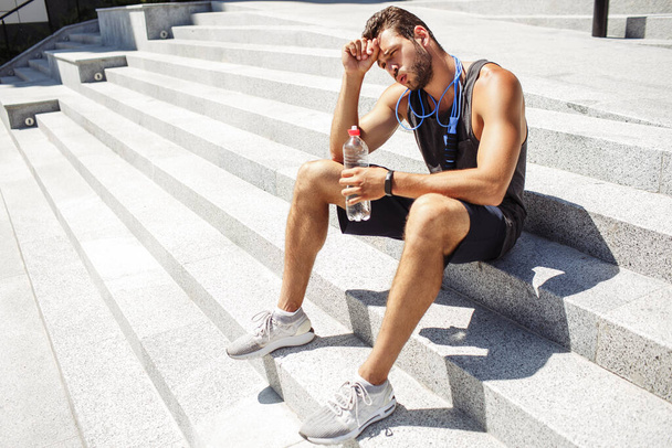 Young man exercising outside. Athletist sit down on steps and resting after workout. Holding water bottle. Having jumping rope around neck. Relax time after exercise. - Photo, Image