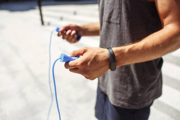 Young man exercising outside. Cut view of athletists hands holding blue jumping rope and ready for exercising. Training outside alone. Sporty guy exercising for better looking and feeling. - Photo, Image