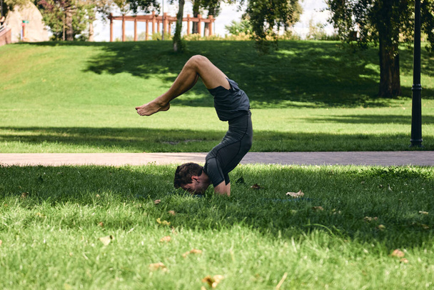 Spor giyimli genç bir adam parkta yoga yapıyor. Asana 'yı dışarıda pratik yap. Yeşil çimlerde yoga minderiyle egzersiz yapan insanlar. Amuda kalkmış, olgun, beyaz bir adam. Güçlü adam zor yoga duruşları deniyor. Fitness ve sağlıklı yaşam tarzı. - Fotoğraf, Görsel