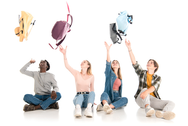 Positive multiethnic teenagers throwing backpacks while sitting on white background - 写真・画像